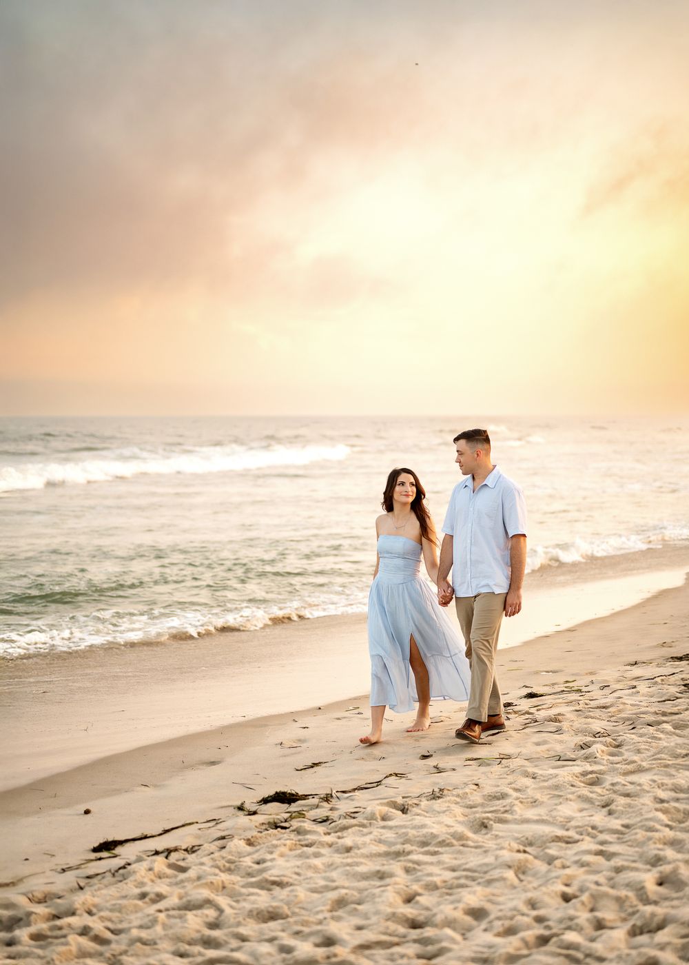 A couple strolls hand in hand along a sandy beach at sunset with gentle waves rolling in beside them.