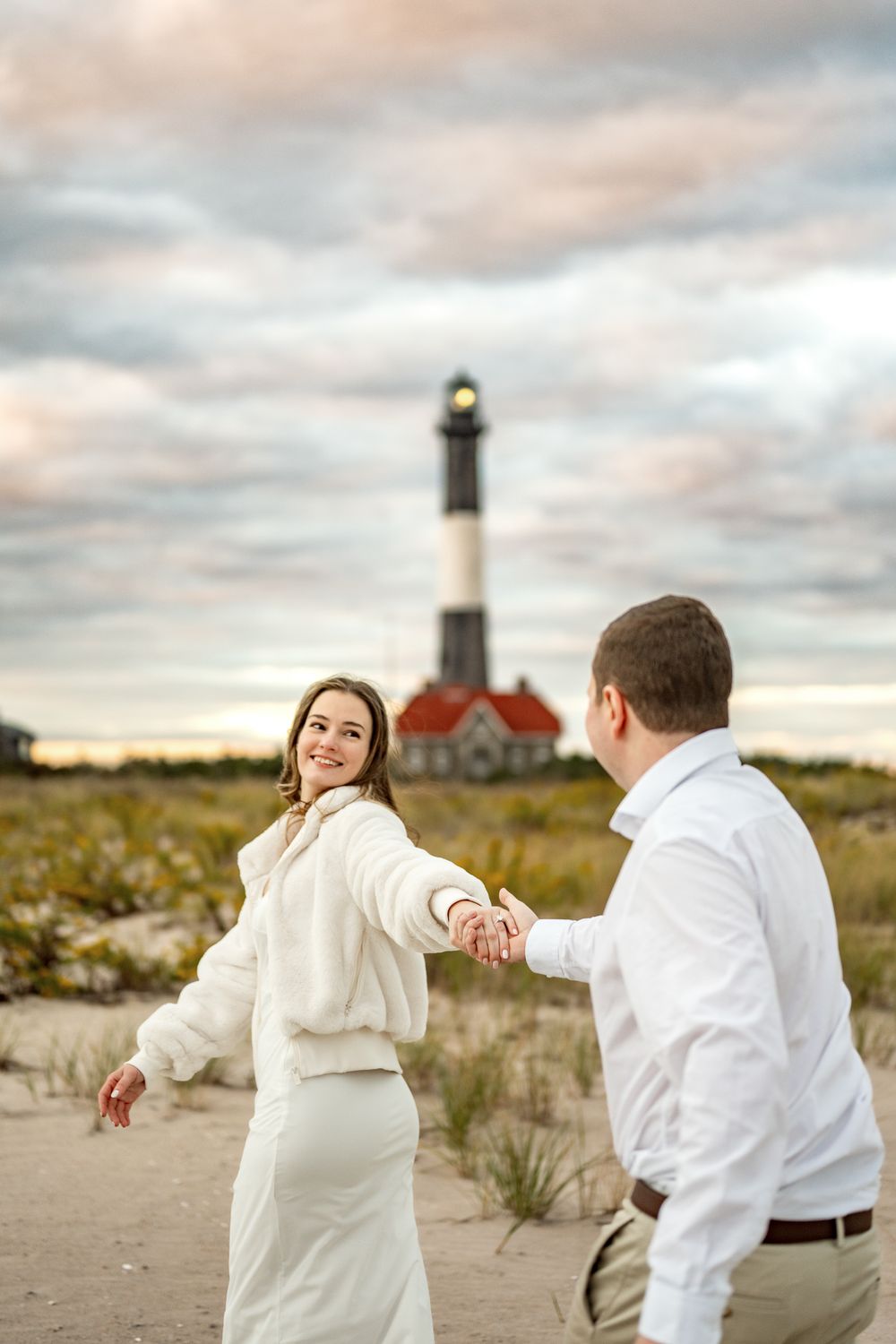 A couple dances playfully on a sandy beach with a lighthouse and red-roofed building in the background.