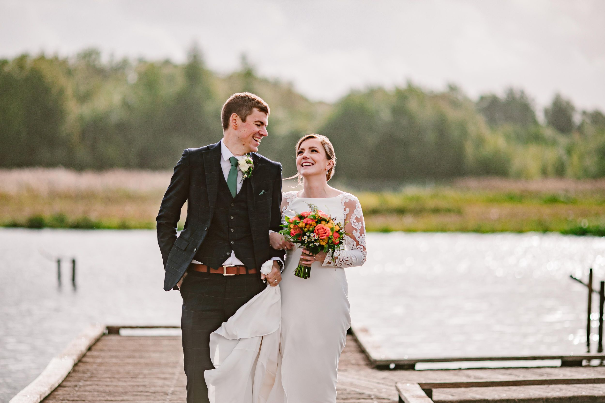beautiful bride looks at her groom walking down the pier at brockholes nature reserve in the background