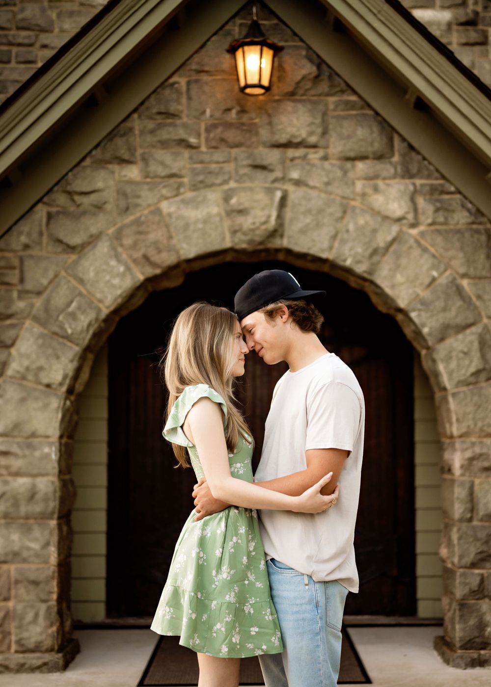 A romantic couple stands together under a stone archway with a glowing lantern above at dusk.