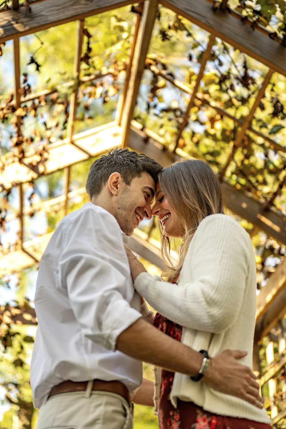 Romantic couple embraces under a sunlit pergola covered in golden autumn leaves.
