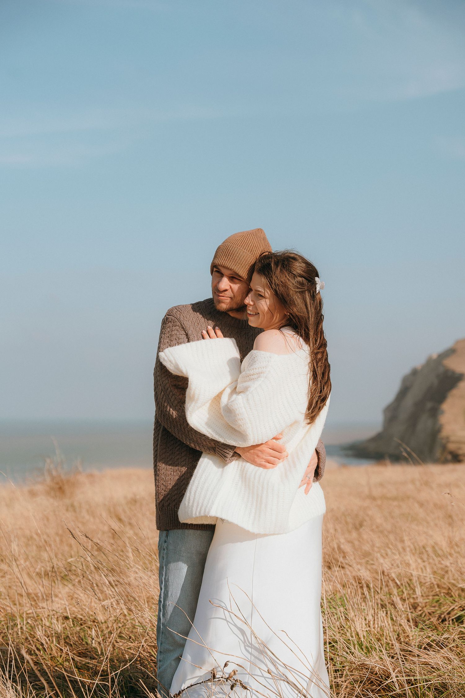A couple embraces in casual winter attire while standing in a golden field by the coast on a sunny day.
