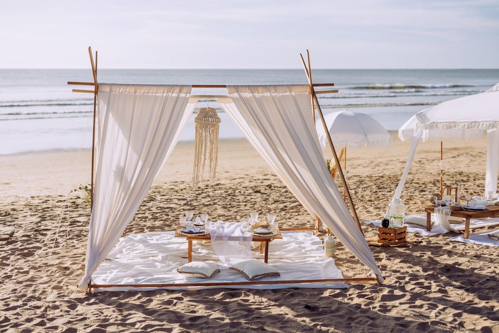 A romantic beach picnic setup with white flowing curtains, pillows and wooden posts creating an intimate cabana on the sand.