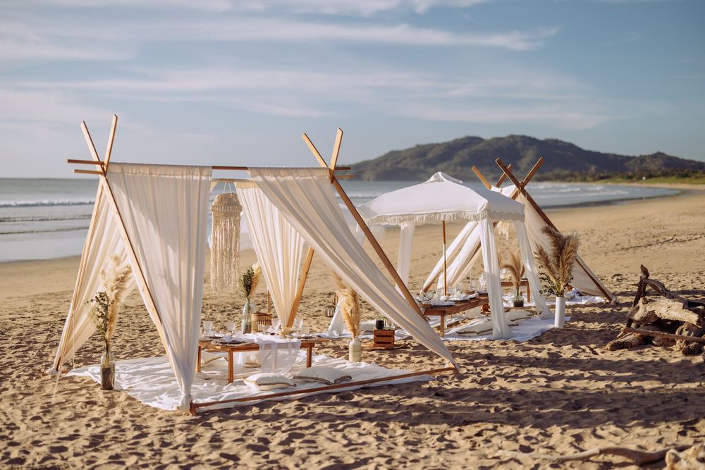 White beach cabanas with wooden posts line a sandy shoreline with mountains in the distance on a sunny day.