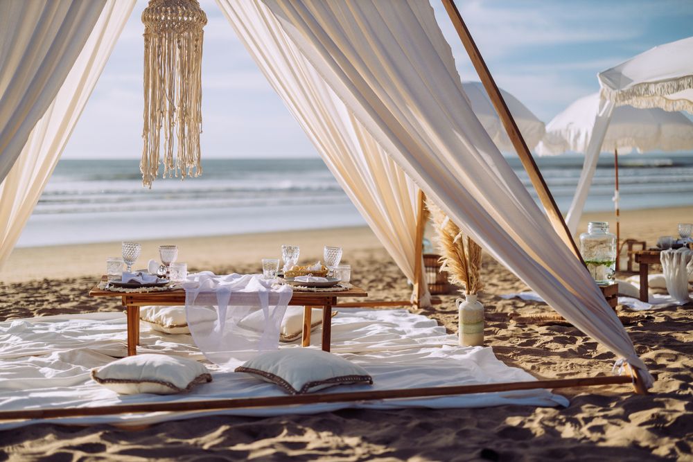 Romantic beach dining setup with flowing white curtains and low tables on the sand by the ocean.