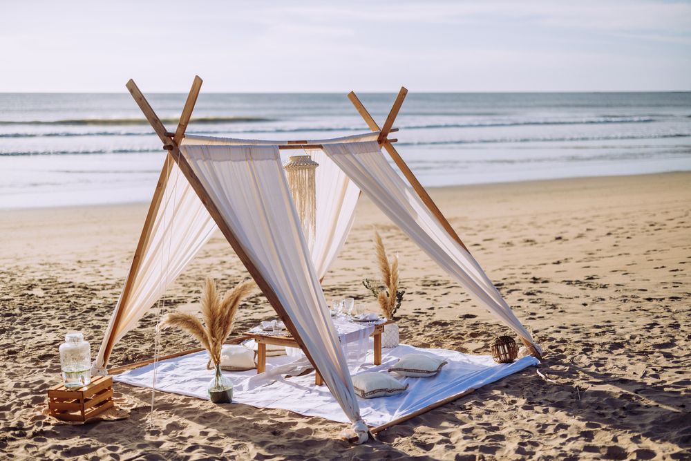 Romantic beach setup with white fabric canopy, wooden poles, and decorative elements on sandy shoreline.