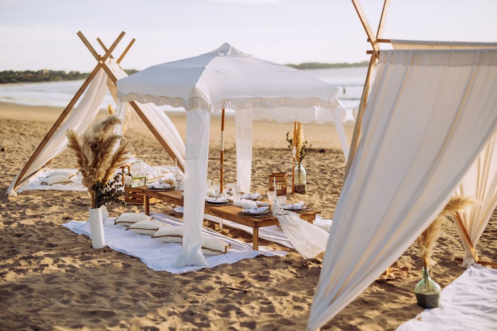 Dreamy beach picnic setup with white fabric canopy, wooden poles, and cushioned seating on the sand at sunset.