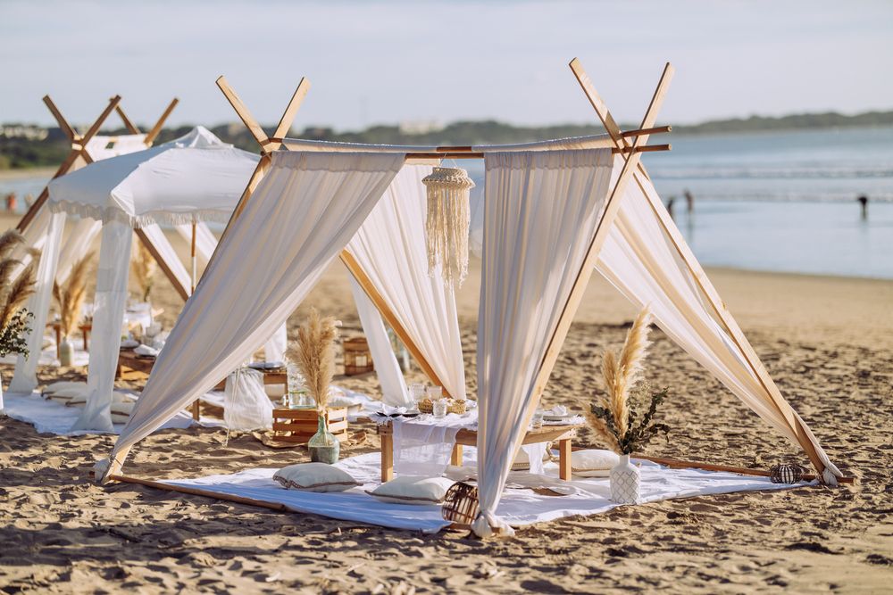 Romantic beach picnic setup with white fabric teepee tents and cushions arranged on the sand by the ocean.