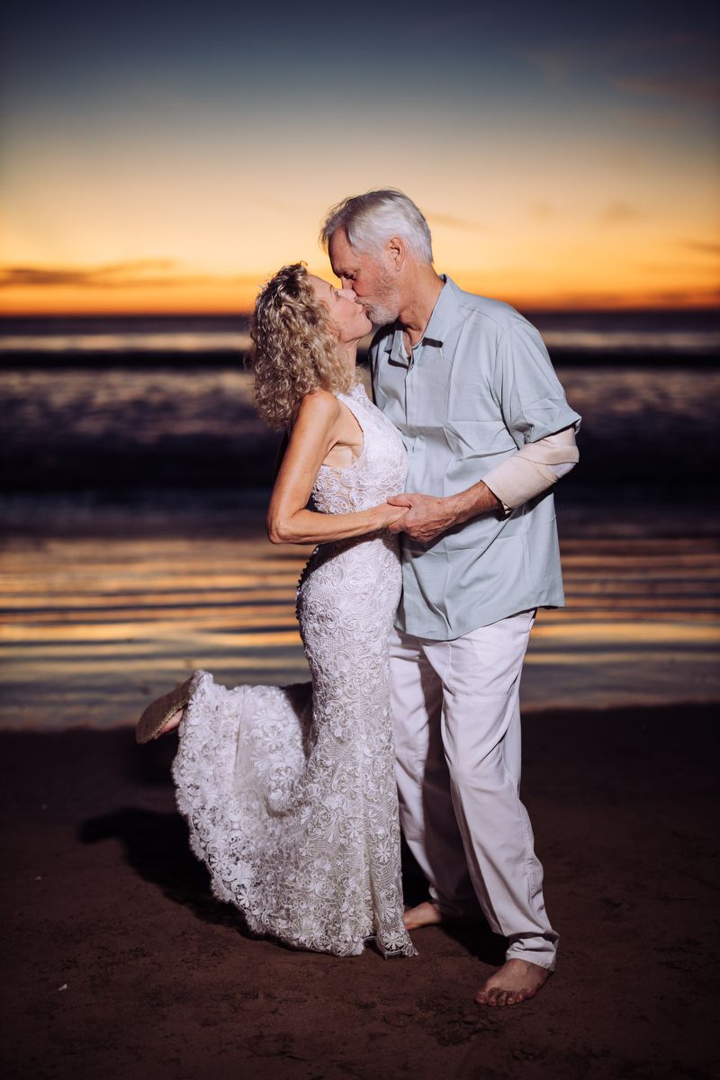 A couple shares a romantic kiss on the beach at sunset while waves lap at the shore.