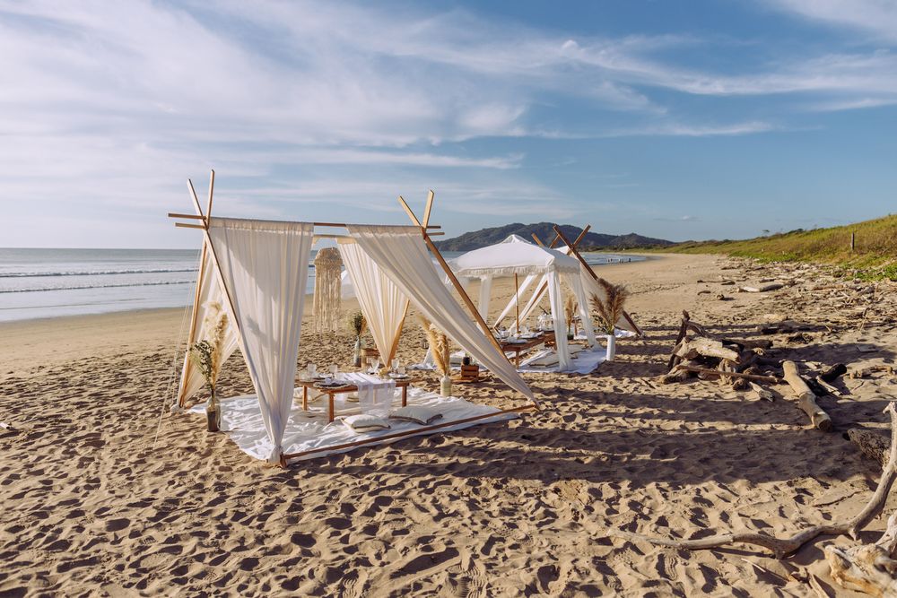 Romantic beach lounge setup with white canopy tents, pillows and blankets on sandy shore during daytime.
