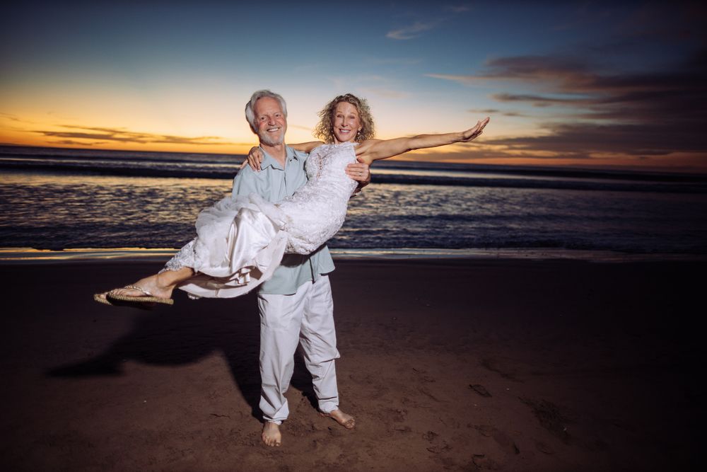 A playful couple on the beach at sunset with someone being carried in white attire against a colorful sky.