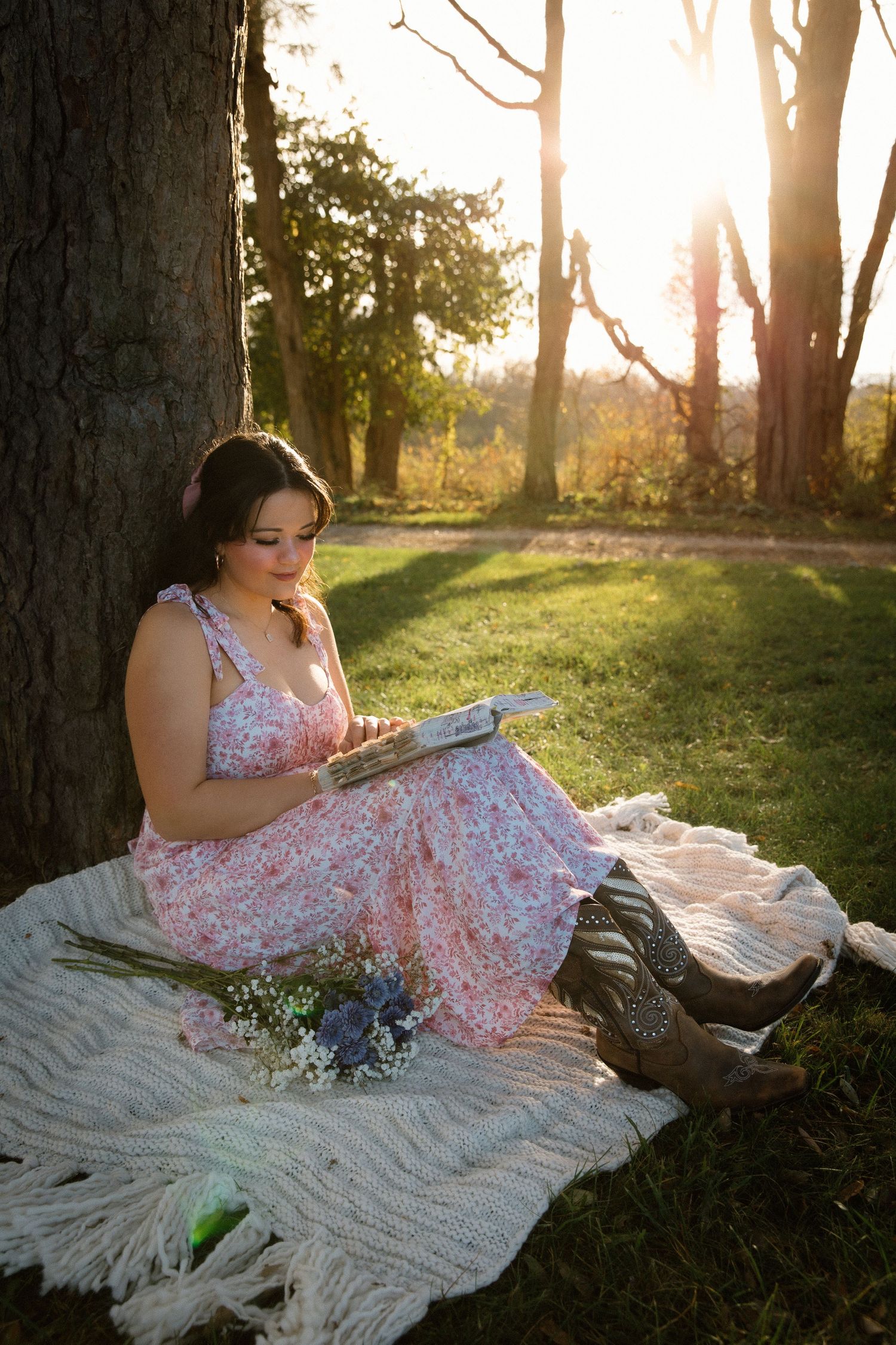 A person in a floral dress reading while sitting on a blanket under a tree at sunset.