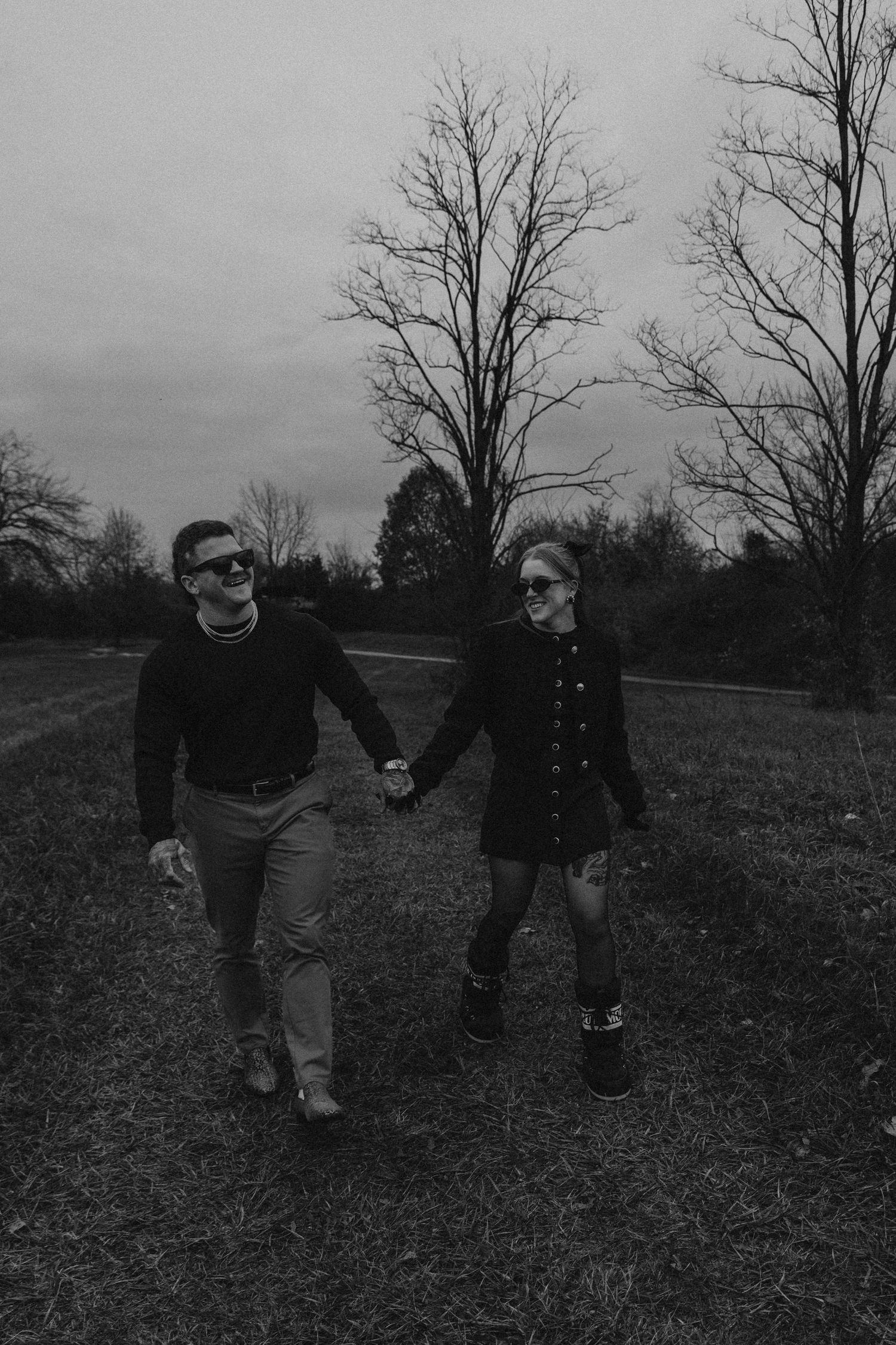 A black and white photo of two people walking hand in hand across an open field with bare trees.