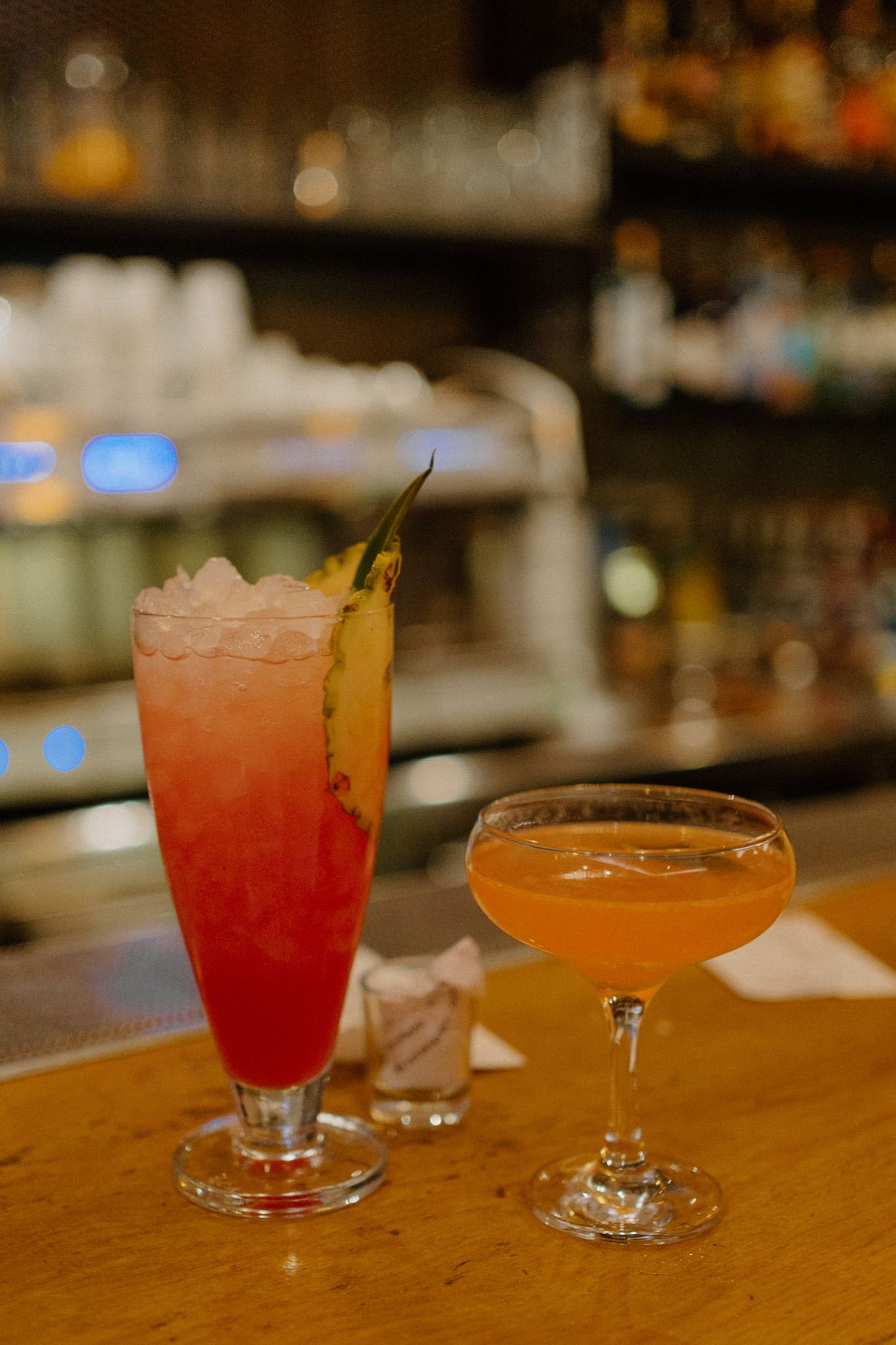 Two craft cocktails on a wooden bar counter, one tall pink drink with pineapple and one orange cocktail in a coupe glass.