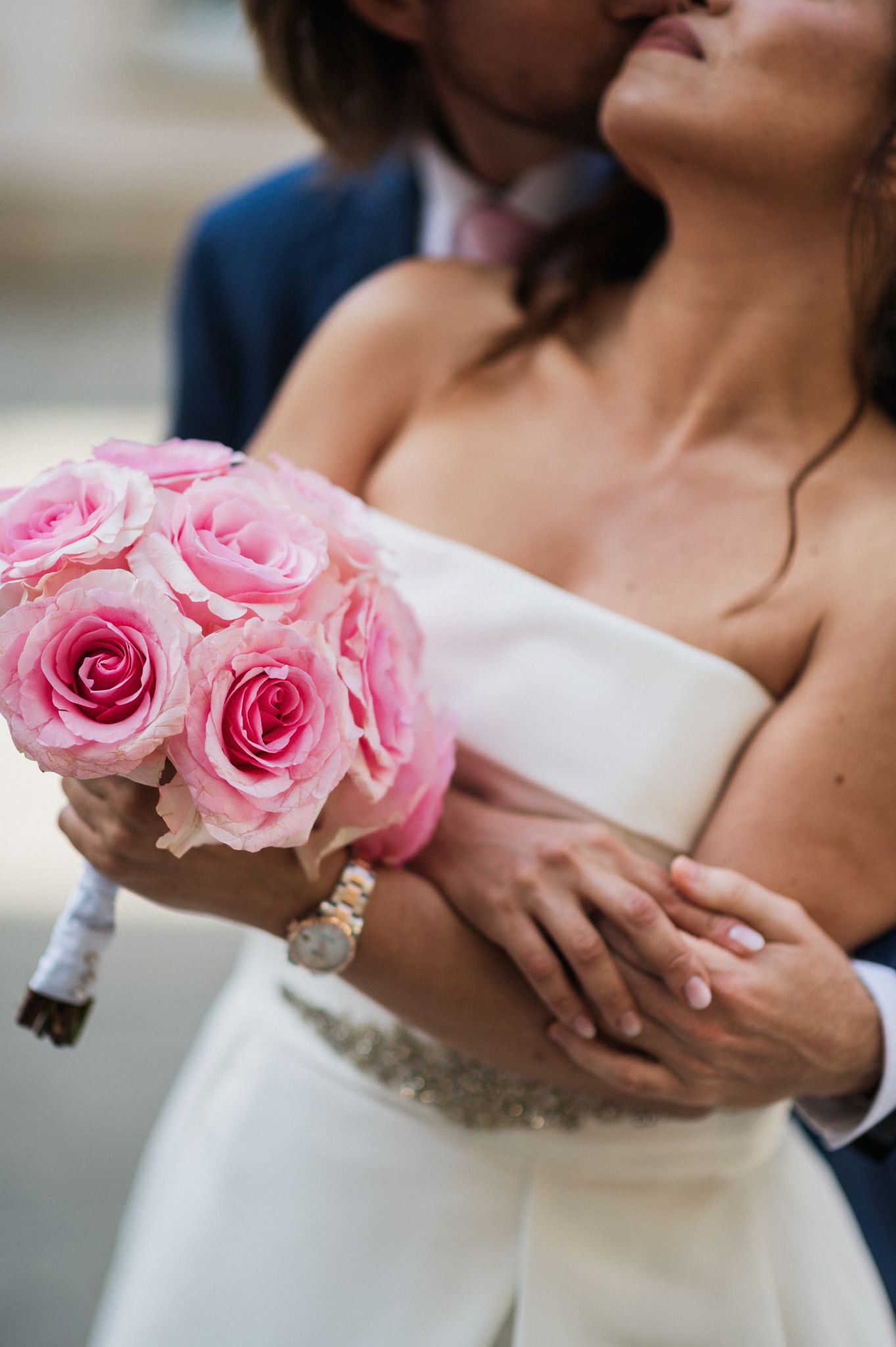 Romantic wedding couple embraces while holding a bouquet of pink roses against a white wedding dress.