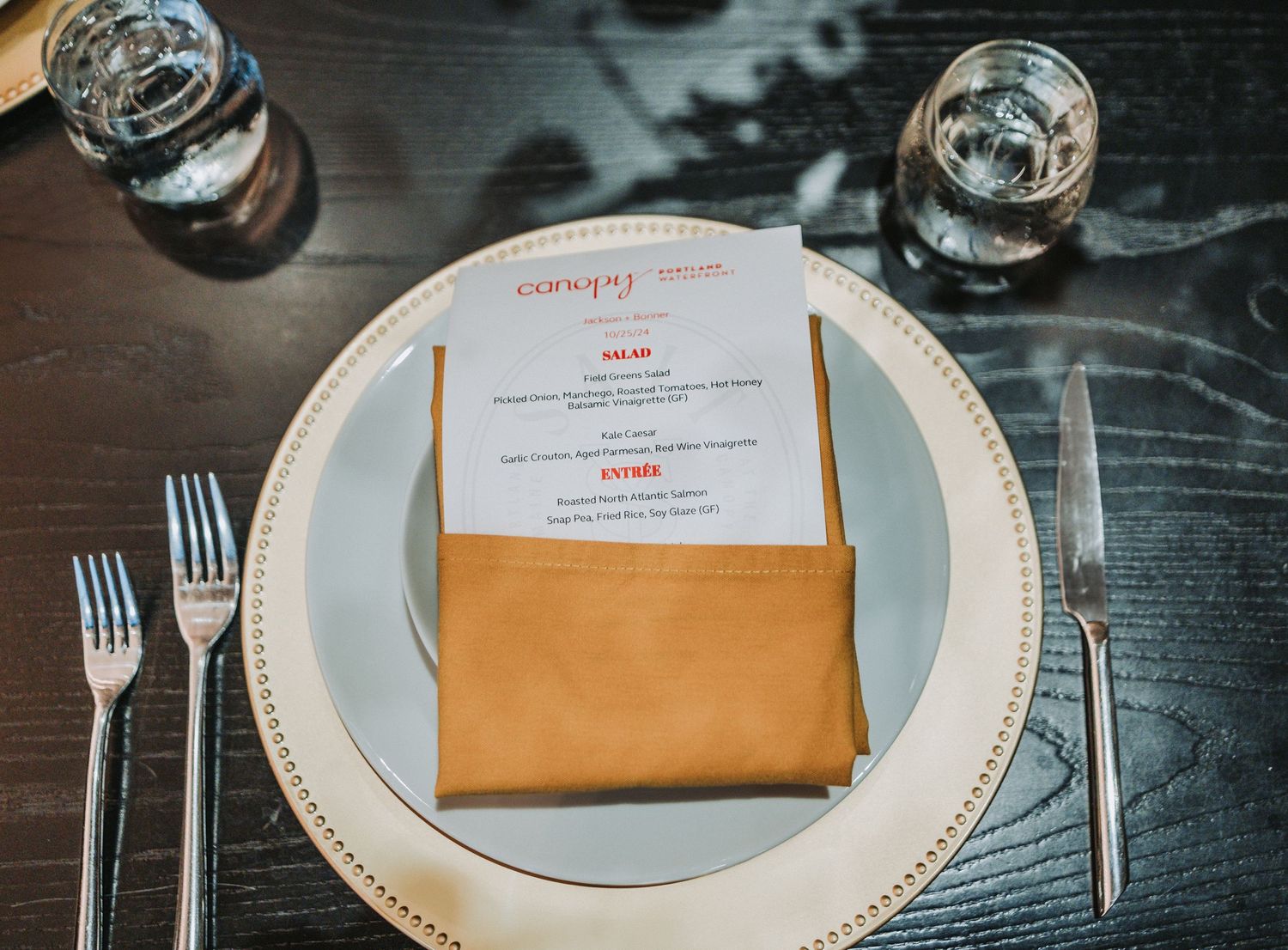 Place setting with white plate, gold napkin holder containing menu, and silverware on dark wooden table with water glasses.  Canopy Hotel.