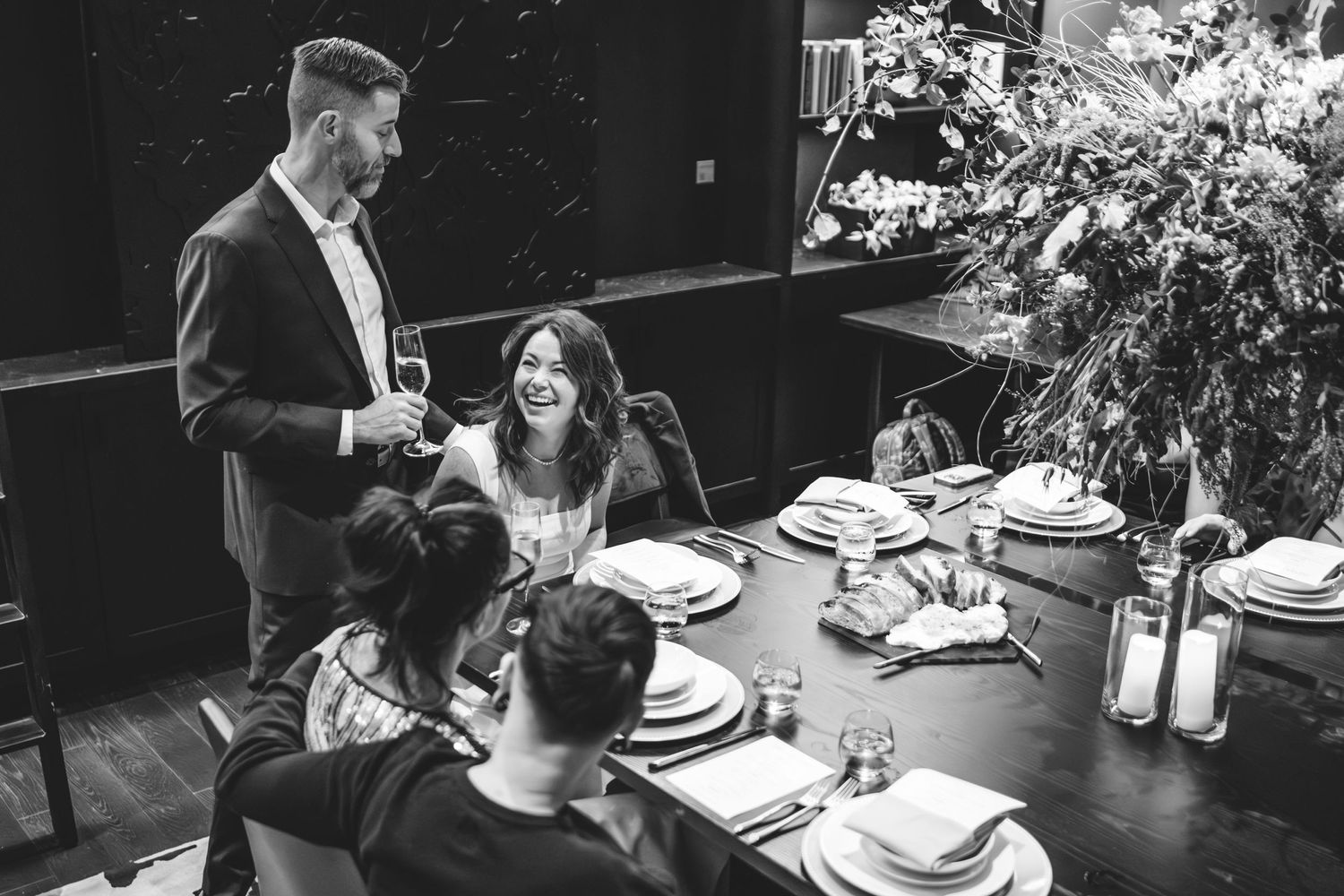 Black and white photo of a bride and groom to be and people gathered around an dinner table with place settings and decorative flowers.
