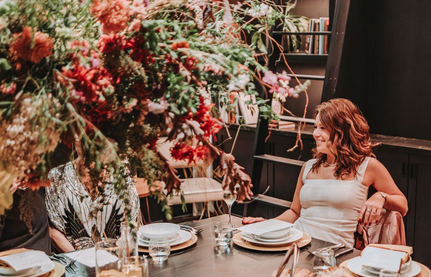 A red floral wedding reception table features elegant place settings and a lush overhead floral arrangement in warm tones.  Canopy Hotel.