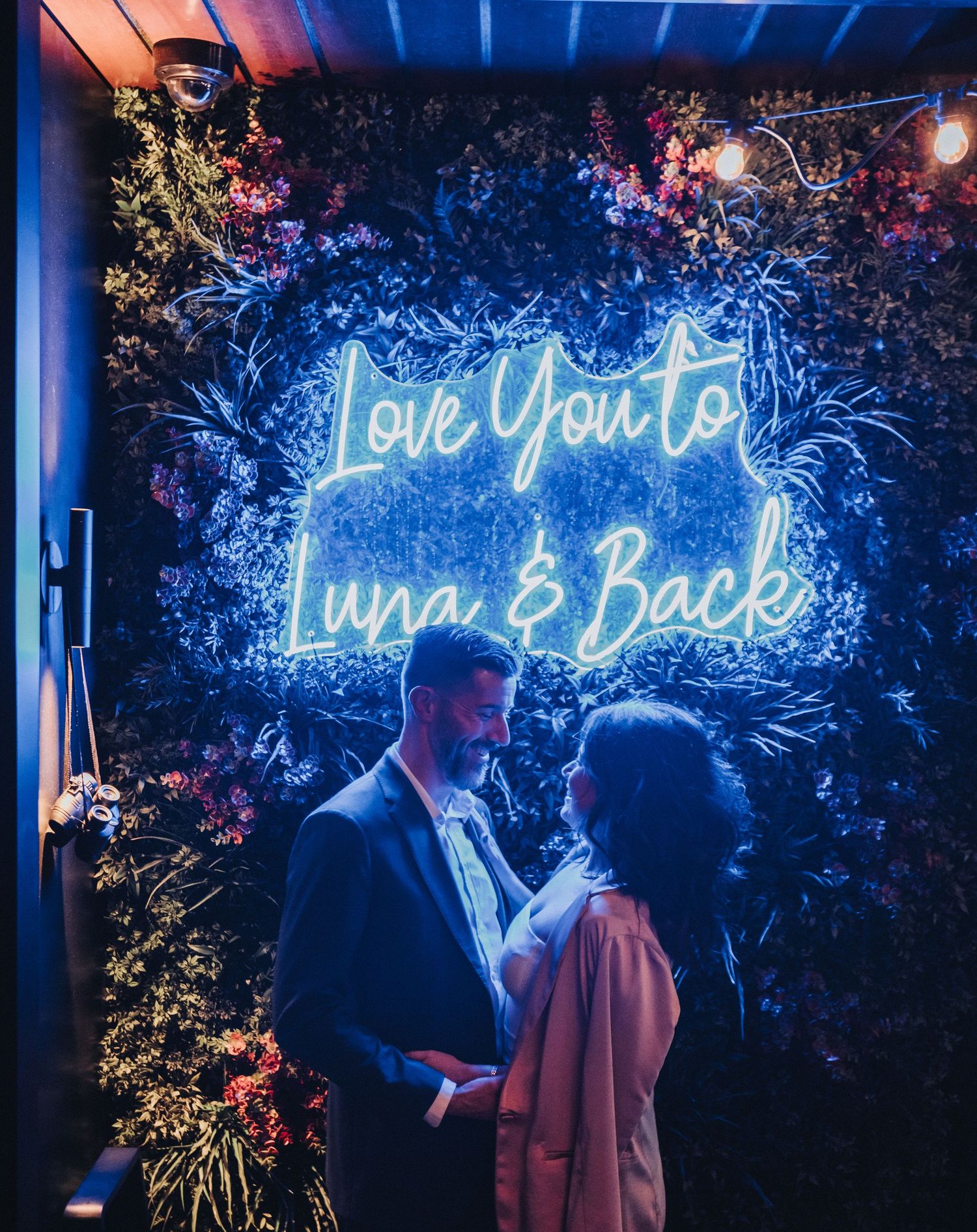 A couple shares a romantic moment under a neon sign reading 'Love You to Luna & Back' with floral decorations.  Located at the Canopy Hotel.
