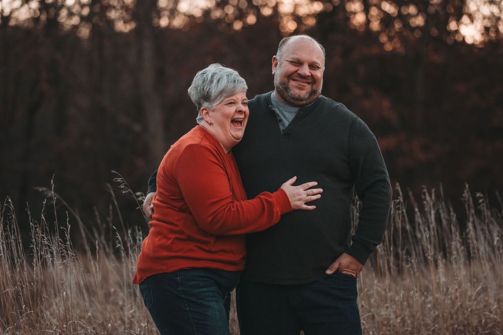 A couple laughs together in a field of tall grass during sunset while wearing casual sweaters.