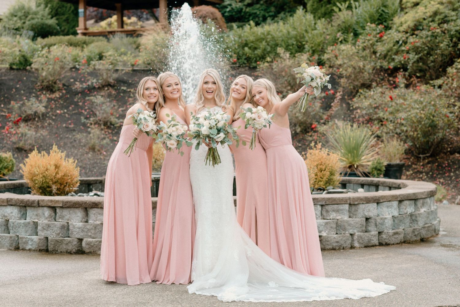 Bridesmaids in pink dresses stand with bride in front of stone wall and fountain.