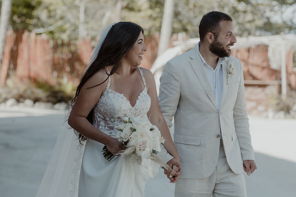 A happy couple holds hands and walks during their beach wedding ceremony with flower bouquet and light colored attire.