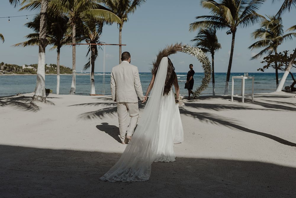 A couple in wedding attire walks along a tropical beach path lined with palm trees and ocean views.