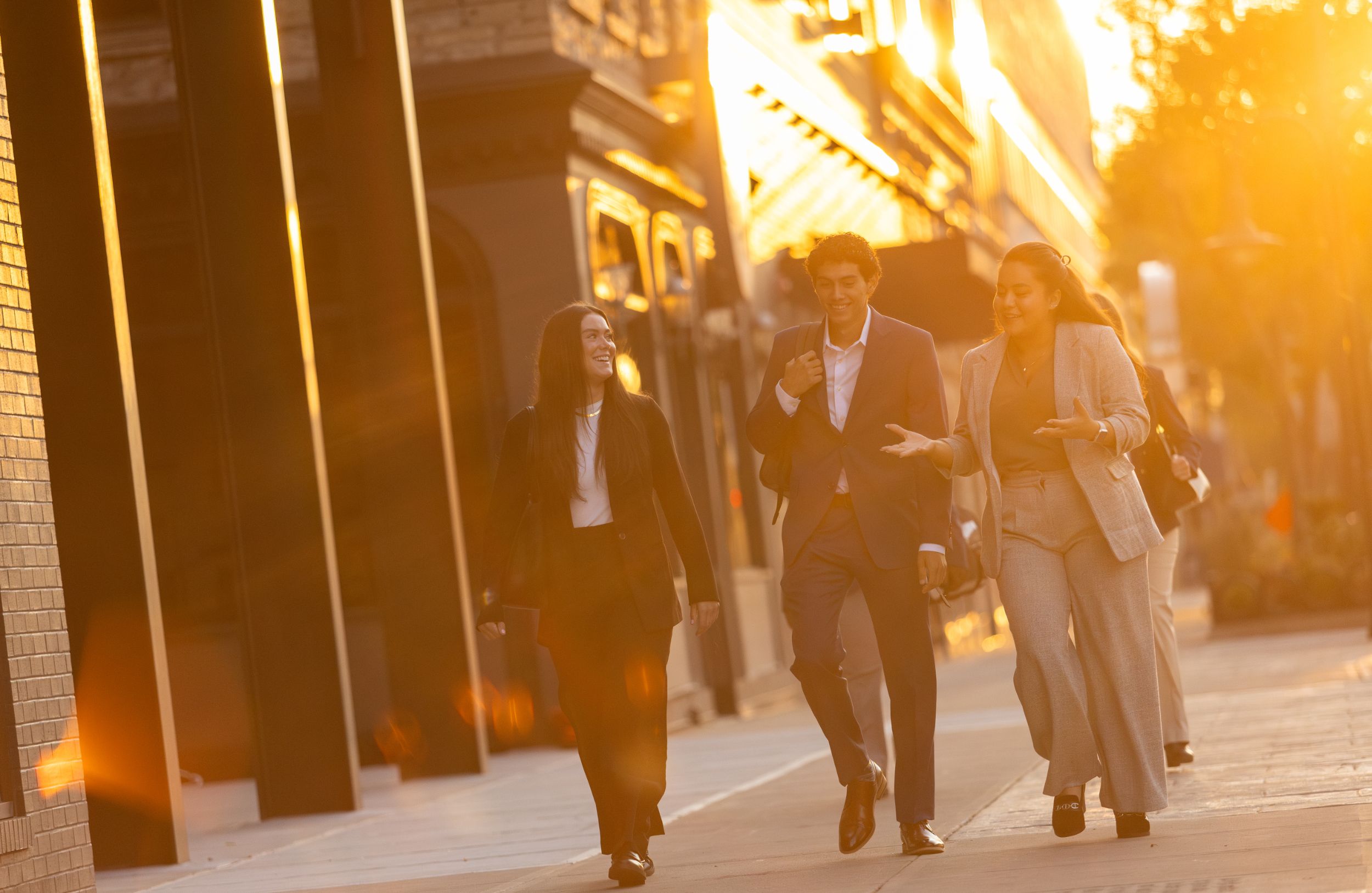 A group of business people walk along a sidewalk in downtown while backlit by the rising sun.