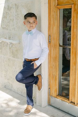 A young boy in a white dress shirt, blue bow tie, navy pants, and tan shoes leans against a stone wall by a wooden door.