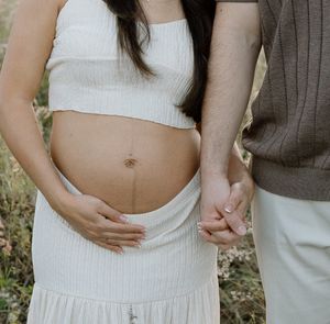 Pregnant woman in white crop top and skirt holding baby bump while partner holds her hand outdoors.