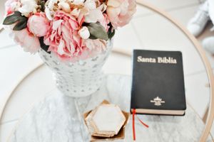A Santa Biblia rests beside a crystal vase with pink peonies on a marble table.