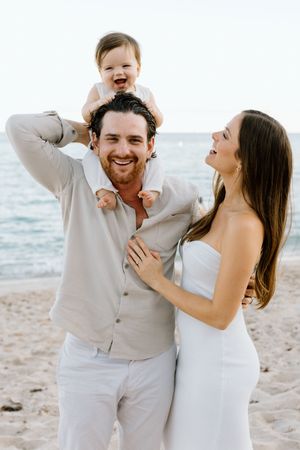 Happy family of three posing on a sandy beach, father holding baby on shoulders with smiling mother in white dress.