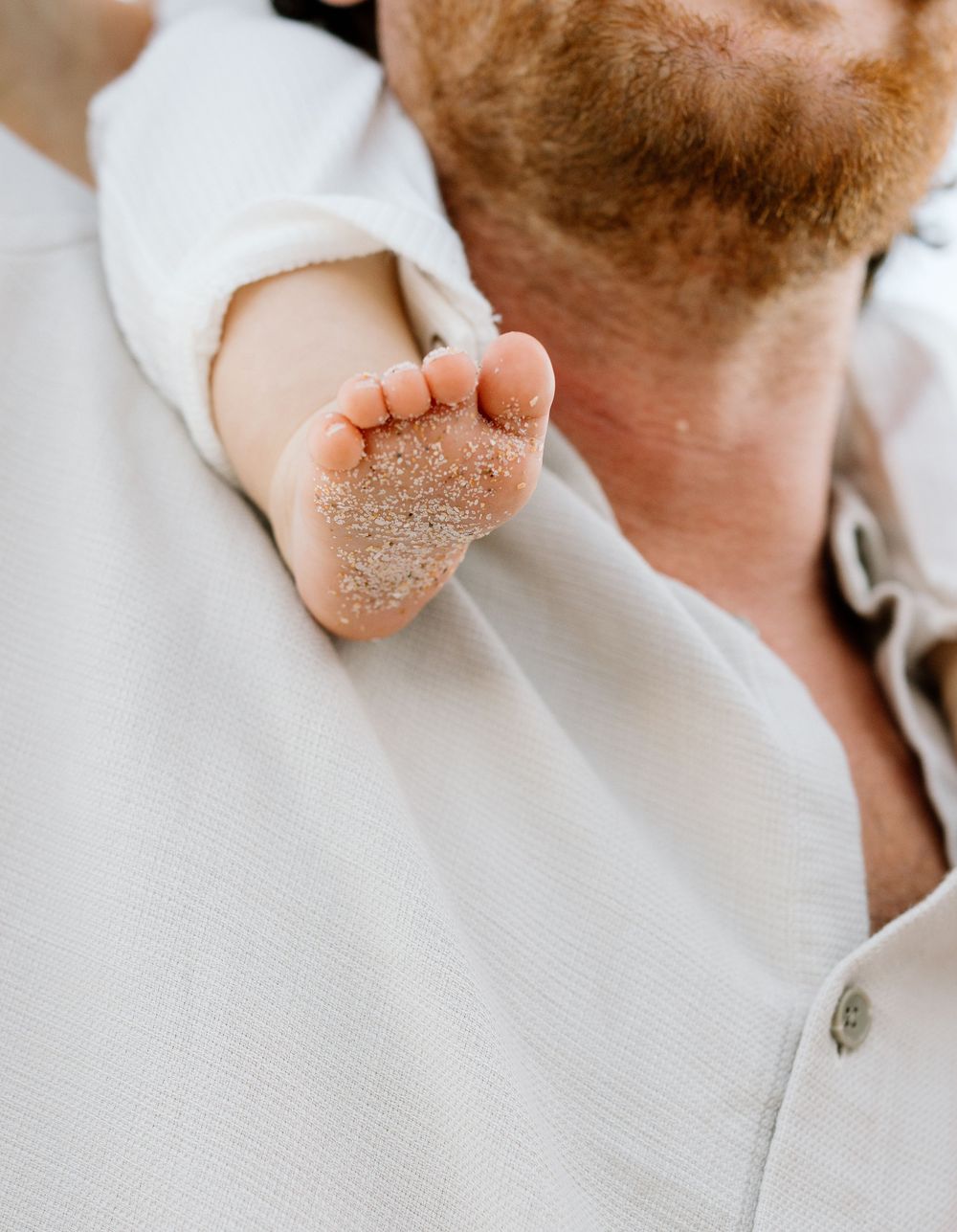 A sandy baby foot resting on a red-bearded man's shoulder in a white shirt.