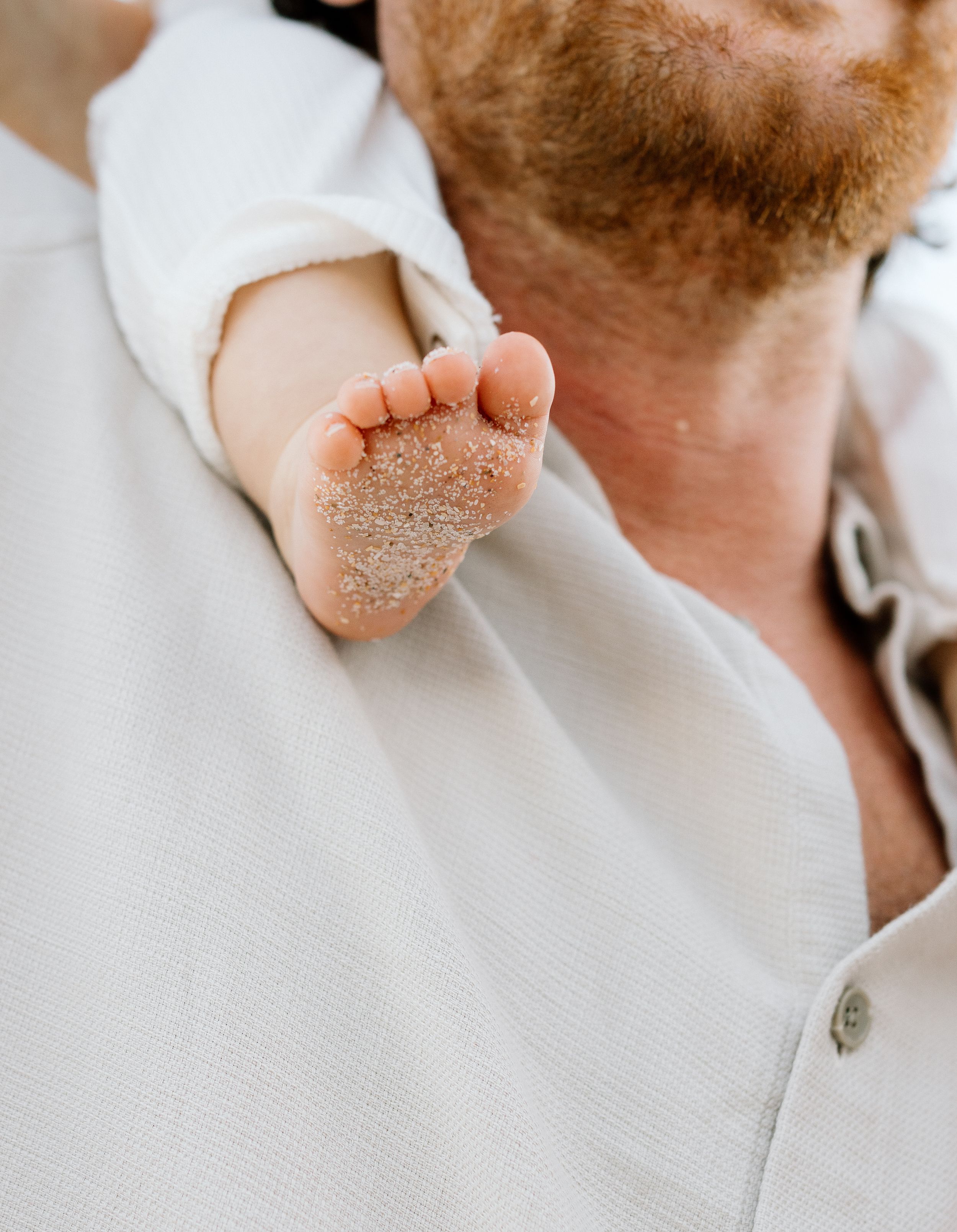 A sandy baby foot resting on a red-bearded man's shoulder in a white shirt.
