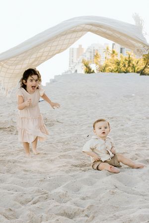Two young children playing on sandy beach under white blanket, girl in pink dress and baby boy sitting in sand.