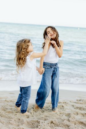 Two young girls laughing and holding hands on a sandy beach, both wearing white tops and jeans.