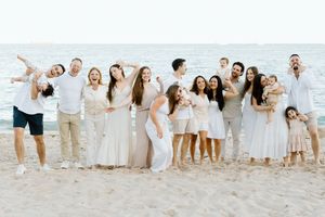 Large family group posing joyfully on a sandy beach wearing coordinated white and beige outfits with ocean in background.