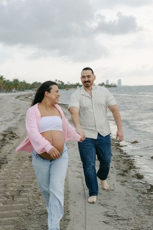 Pregnant couple holding hands and walking along a sandy beach shoreline, woman in pink cardigan and white crop top.