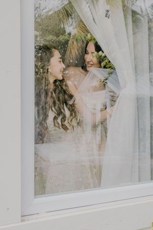 Two brides smiling and embracing behind a sheer curtained window on their wedding day.