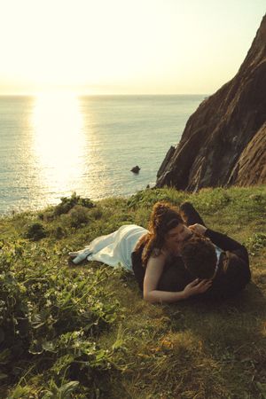 A couple rests on a grassy cliff overlooking the ocean at sunset, with golden light reflecting on the calm sea below.