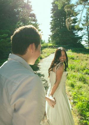 Couple holding hands on a sunny forest path, woman in white dress smiling joyfully at her partner.