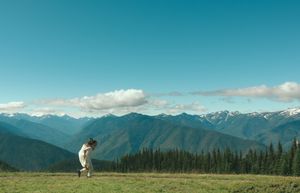 Person standing on a green meadow with panoramic mountain views under a bright blue sky with scattered clouds.