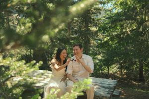 Couple sitting on wooden deck in sunlit forest, laughing and sharing a joyful moment surrounded by green pine trees.