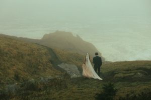 Two figures stand atop a misty mountain cliff overlooking a dramatic coastal landscape in moody weather.