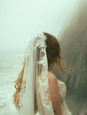 A bride in a delicate lace veil and gown is photographed in dreamy light with flowing long hair.