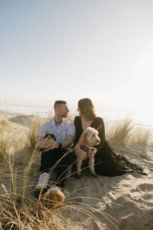 A couple sits on a sandy beach at sunset with their small dog among tall beach grass by the ocean.
