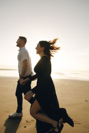 A couple runs together along a sunset beach while their hair and clothing blow in the wind.