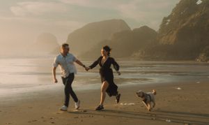 Couple joyfully running on a misty beach at sunset with their dog against a backdrop of coastal cliffs.