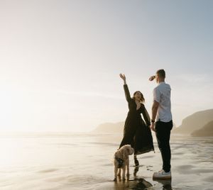 Couple and their dog standing on beach at sunset with person raising arm in joyful pose.