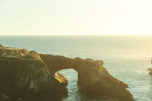 A natural stone arch formation along the coastline at sunset with silhouettes visible on top against golden light.